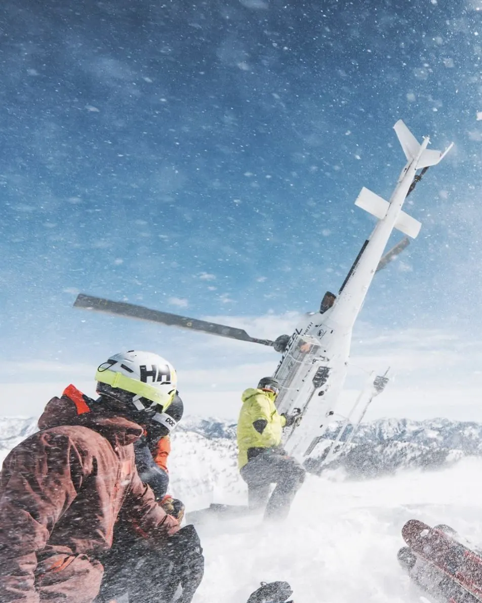 Freeride skiers crouching in the snow as a helicopter lifts off from an alpine summit