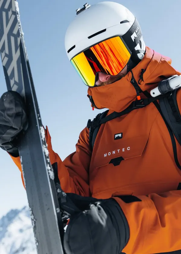 Skier in an orange ski jacket, white helmet and mirrored goggles, holding a pair of skis